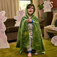 A child wearing a green Dragon’s Tale Velour Cape by Magnetic Me stands smiling indoors, surrounded by large animal cutouts and soft furniture in a cozy room—the perfect setting for imaginative costumes and pretend play.