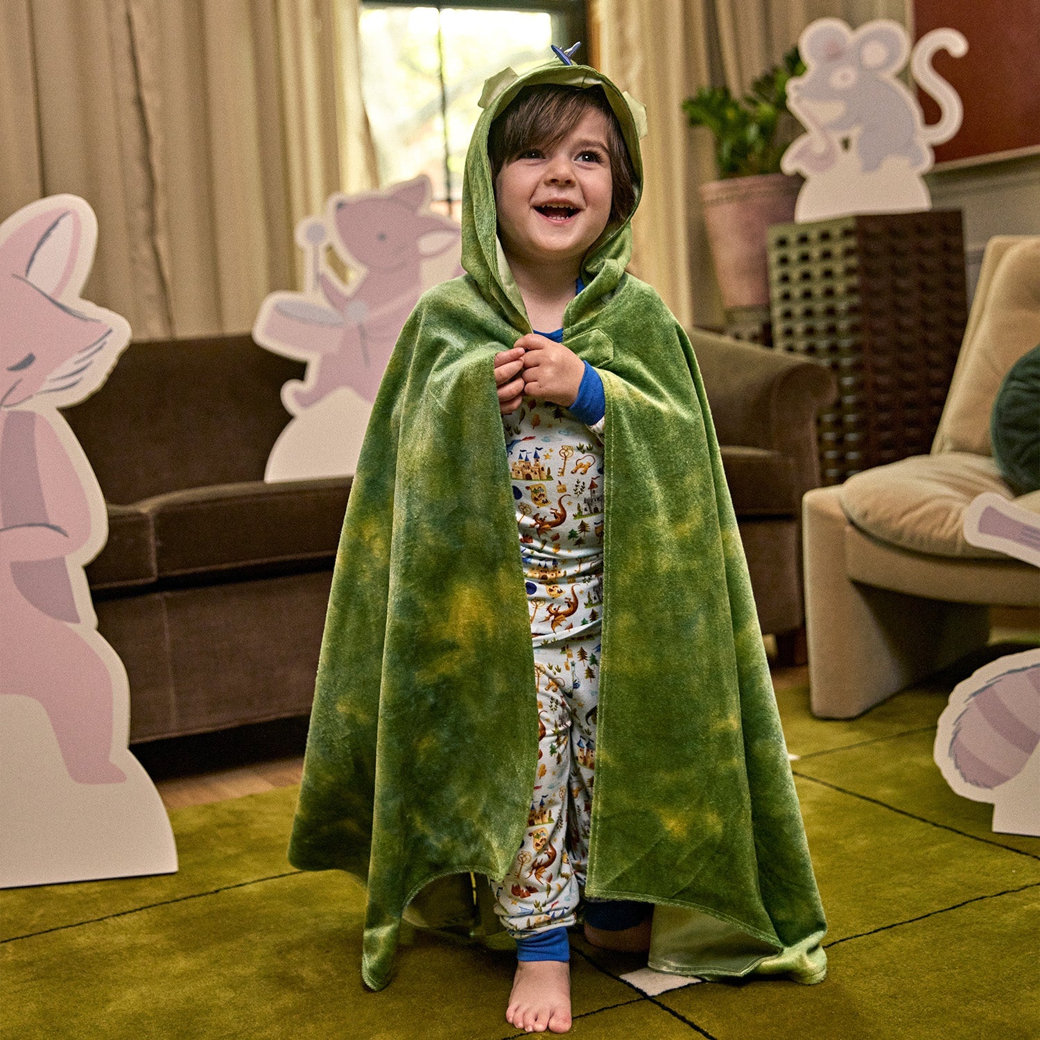 A child wearing a green Dragon’s Tale Velour Cape by Magnetic Me stands smiling indoors, surrounded by large animal cutouts and soft furniture in a cozy room—the perfect setting for imaginative costumes and pretend play.