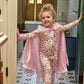 A young child wearing patterned pajamas and a pink Fairytale Era Tulle Cape by Magnetic Me stands in a tiled room doorway, smiling and posing with one hand on the door handle—ready for pretend play adventures.