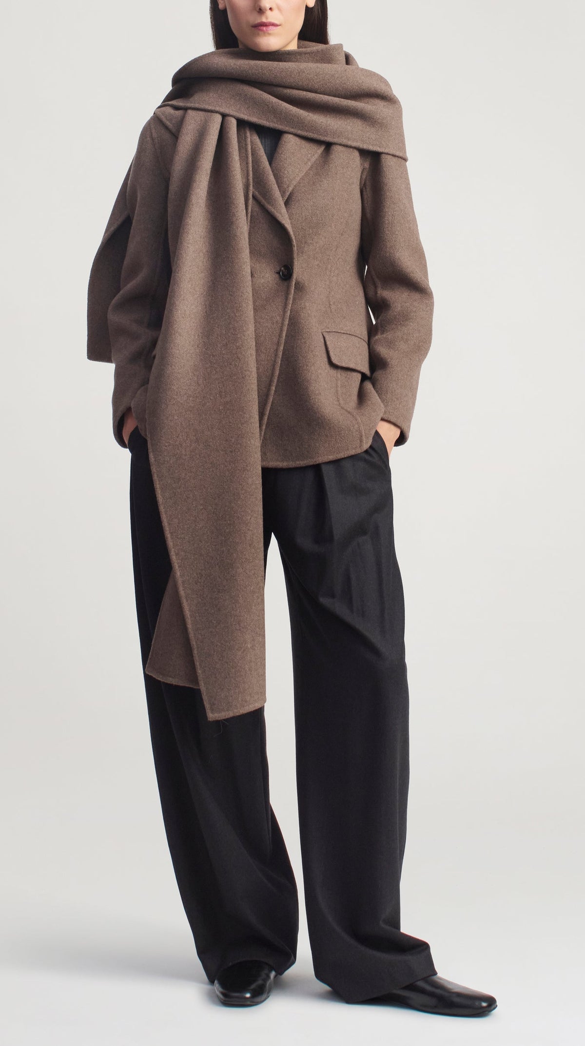 A person wearing a Kallmeyer Sablier Scarf Blazer in brown virgin wool with an integrated scarf draped over the shoulders, paired with loose black trousers.