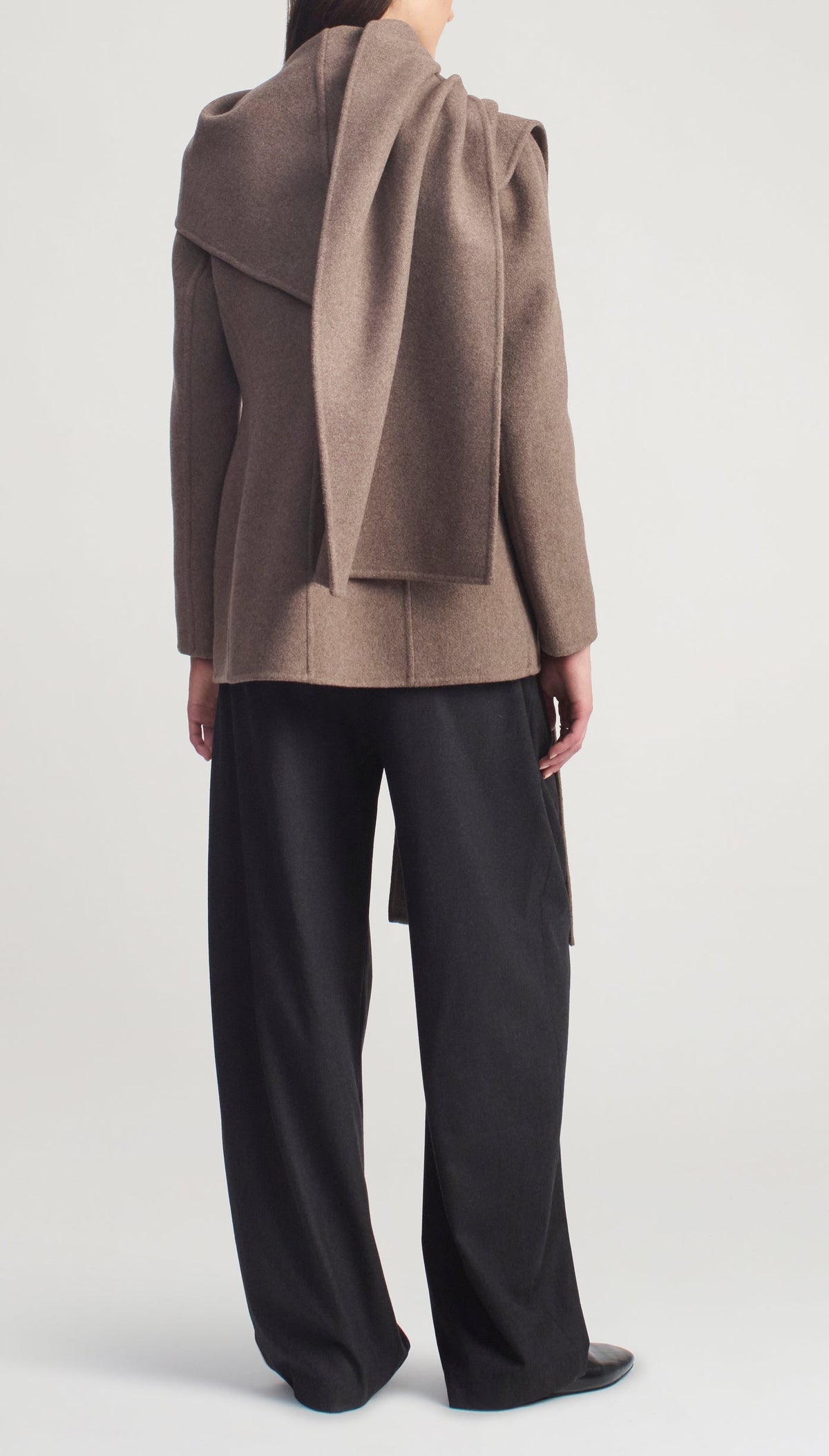 Back view of a person wearing a brown virgin wool Kallmeyer Sablier Scarf Blazer with an integrated scarf draped over the shoulder, paired with black wide-leg trousers.