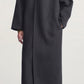 A person wearing the Kallmeyer Dorothy Robe Coat, a long, architectural piece in dark gray with an oversized shawl collar, paired with black stockings and black low-heeled shoes, stands against a plain light background.