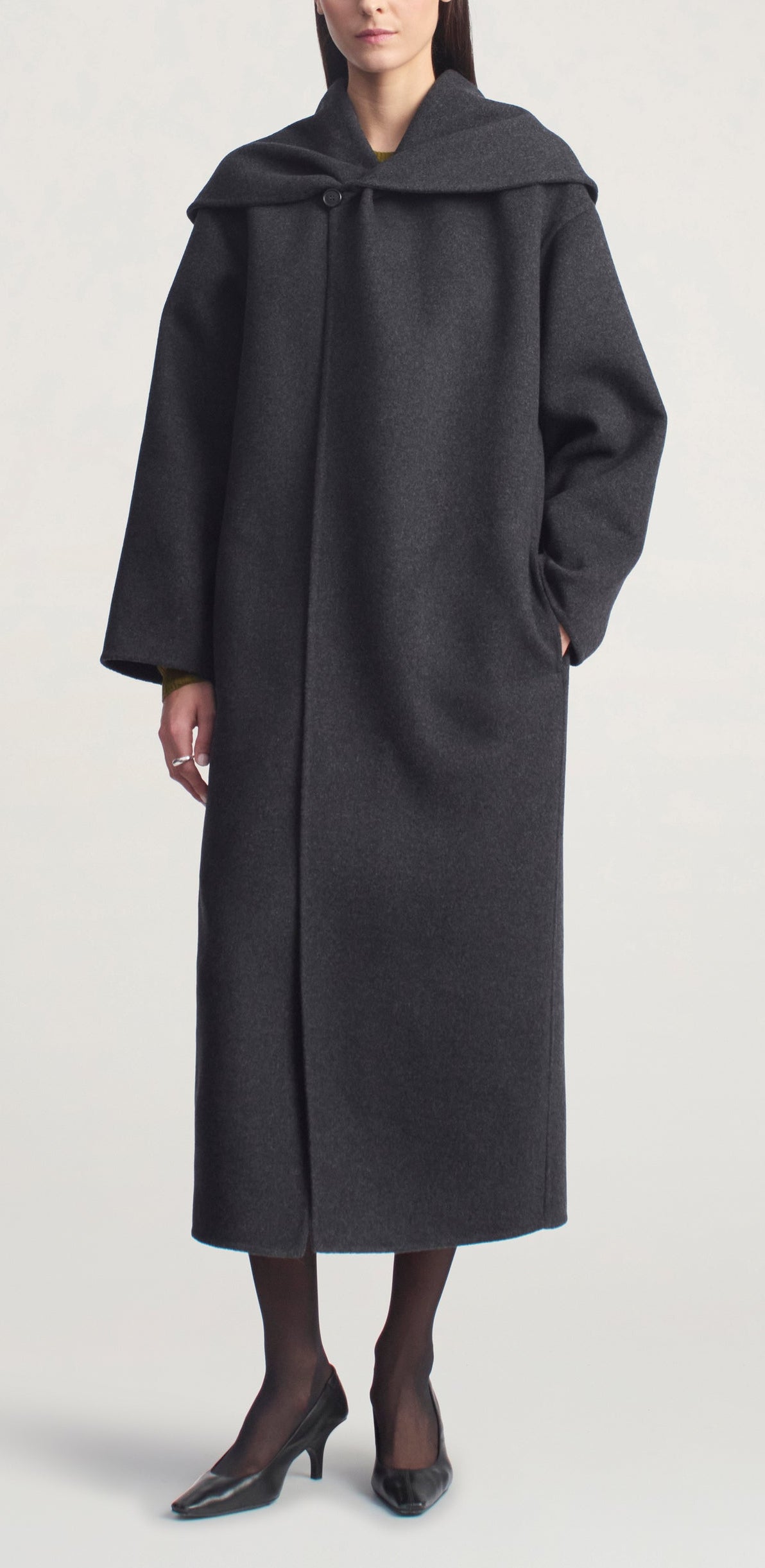 A person wearing the Kallmeyer Dorothy Robe Coat, a long, architectural piece in dark gray with an oversized shawl collar, paired with black stockings and black low-heeled shoes, stands against a plain light background.