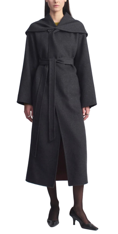 A woman stands against a white background wearing the Kallmeyer Dorothy Robe Coat—a long, dark gray architectural piece crafted from charcoal virgin wool with an oversized shawl collar and wide sleeves—paired with black tights and black heels.