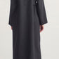 A woman stands facing away, wearing the Kallmeyer Dorothy Robe Coat in long charcoal virgin wool with an oversized shawl collar, paired with black tights and black shoes against a plain background.