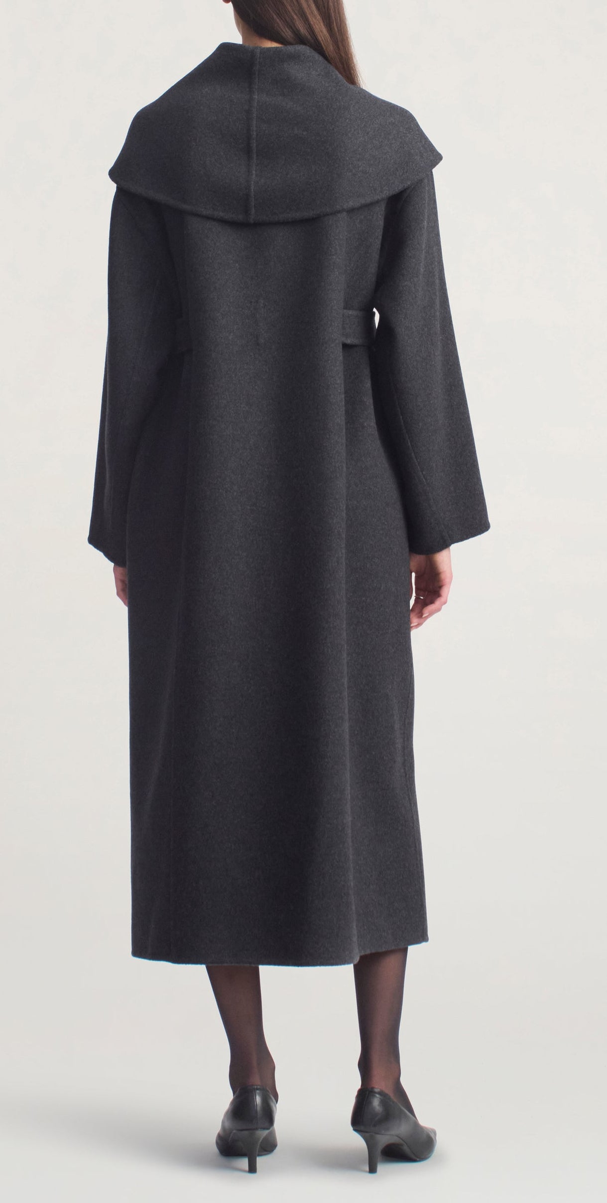 A woman stands facing away, wearing the Kallmeyer Dorothy Robe Coat in long charcoal virgin wool with an oversized shawl collar, paired with black tights and black shoes against a plain background.