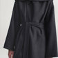 A person stands against a plain light background wearing the Kallmeyer Dorothy Robe Coat, an architectural piece crafted from charcoal virgin wool with a dark gray belted silhouette, wide sleeves, and an oversized shawl collar.
