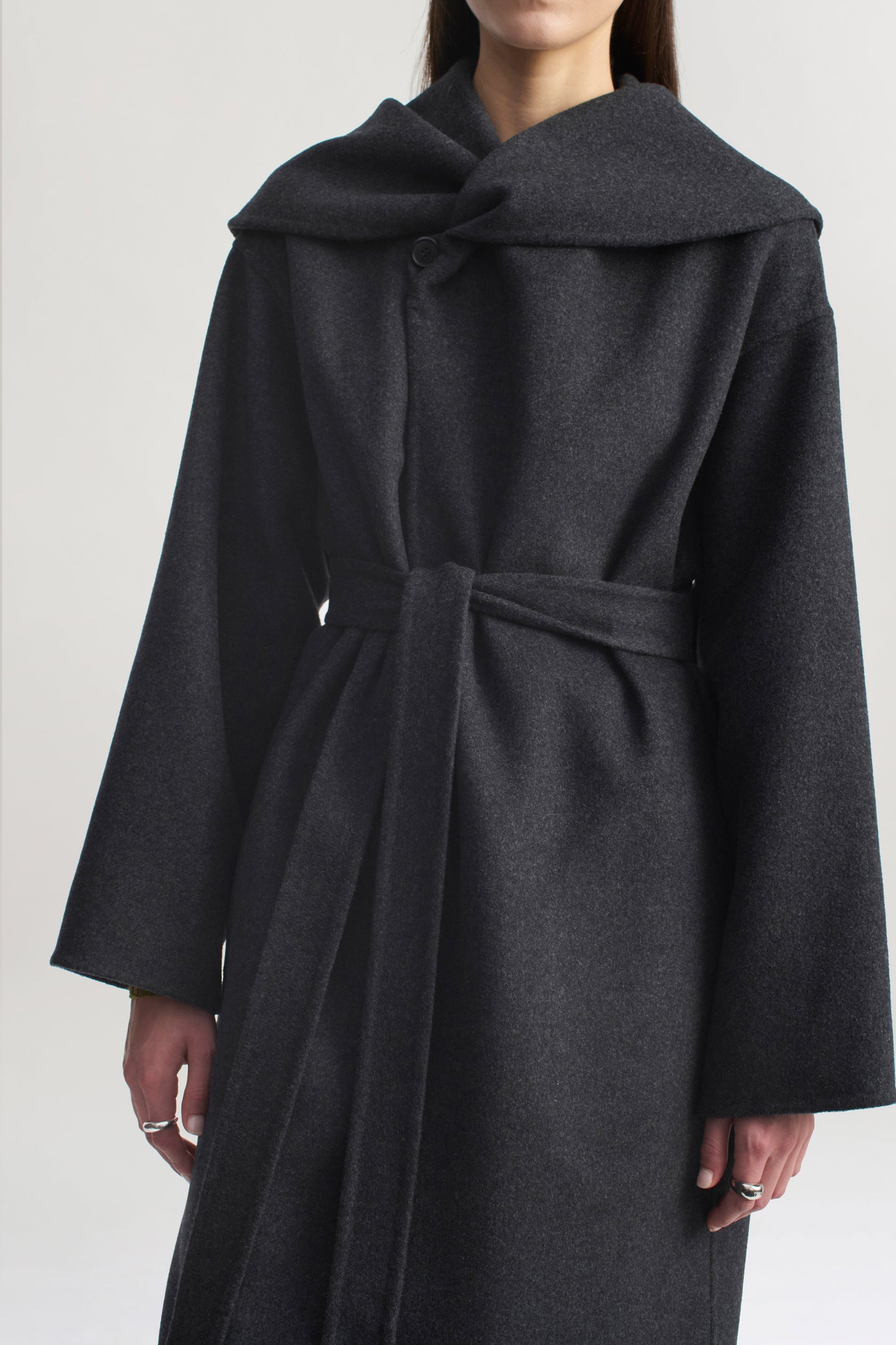 A person stands against a plain light background wearing the Kallmeyer Dorothy Robe Coat, an architectural piece crafted from charcoal virgin wool with a dark gray belted silhouette, wide sleeves, and an oversized shawl collar.