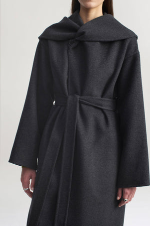A person stands against a plain light background wearing the Kallmeyer Dorothy Robe Coat, an architectural piece crafted from charcoal virgin wool with a dark gray belted silhouette, wide sleeves, and an oversized shawl collar.