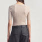 A person stands facing away, wearing the Kallmeyer Pika Short Sleeve V-Neck in beige, paired with high-waisted dark gray trousers featuring belt loops and pocket flaps.