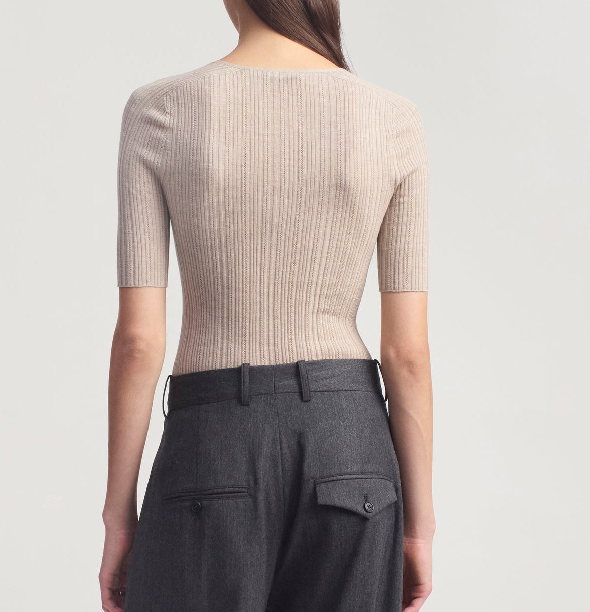 A person stands facing away, wearing the Kallmeyer Pika Short Sleeve V-Neck in beige, paired with high-waisted dark gray trousers featuring belt loops and pocket flaps.