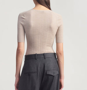 A person stands facing away, wearing the Kallmeyer Pika Short Sleeve V-Neck in beige, paired with high-waisted dark gray trousers featuring belt loops and pocket flaps.