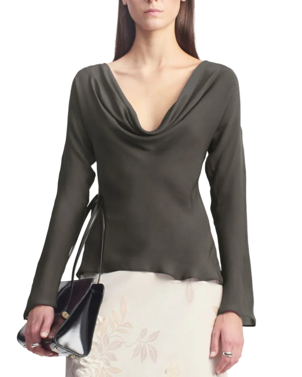 A woman stands against a white background, dressed in Kallmeyer’s Wilka Blouse—an elegant dark, long-sleeved cowl neck top crafted from luxurious silk georgette. She pairs it with a light floral skirt and carries a black handbag. This sophisticated look is made in New York.