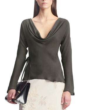 A woman stands against a white background, dressed in Kallmeyer’s Wilka Blouse—an elegant dark, long-sleeved cowl neck top crafted from luxurious silk georgette. She pairs it with a light floral skirt and carries a black handbag. This sophisticated look is made in New York.