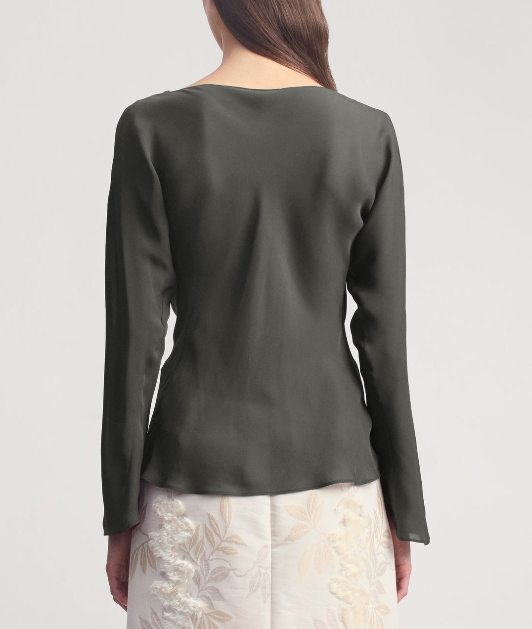 A woman stands facing away, wearing the Wilka Blouse by Kallmeyer—a long-sleeve cowl neck blouse crafted from luxurious silk georgette—and a white skirt with a subtle floral pattern, both thoughtfully made in New York.