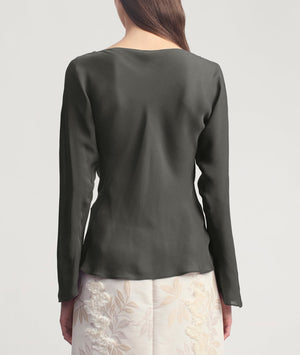 A woman stands facing away, wearing the Wilka Blouse by Kallmeyer—a long-sleeve cowl neck blouse crafted from luxurious silk georgette—and a white skirt with a subtle floral pattern, both thoughtfully made in New York.
