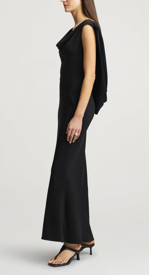Side profile of a person in a black viscose cady Gilda Bias Dress with a cowl neckline, featuring an attached draped back panel and black heeled sandals.