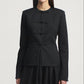 A woman wears the Tilda Jacket by Kallmeyer—a structured, collarless slim jacket in black Grampians Twill with four horizontal bows on the front—paired with a matching black skirt and featuring a subtle peplum silhouette, set against a plain light background.