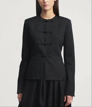 A woman wears the Tilda Jacket by Kallmeyer—a structured, collarless slim jacket in black Grampians Twill with four horizontal bows on the front—paired with a matching black skirt and featuring a subtle peplum silhouette, set against a plain light background.