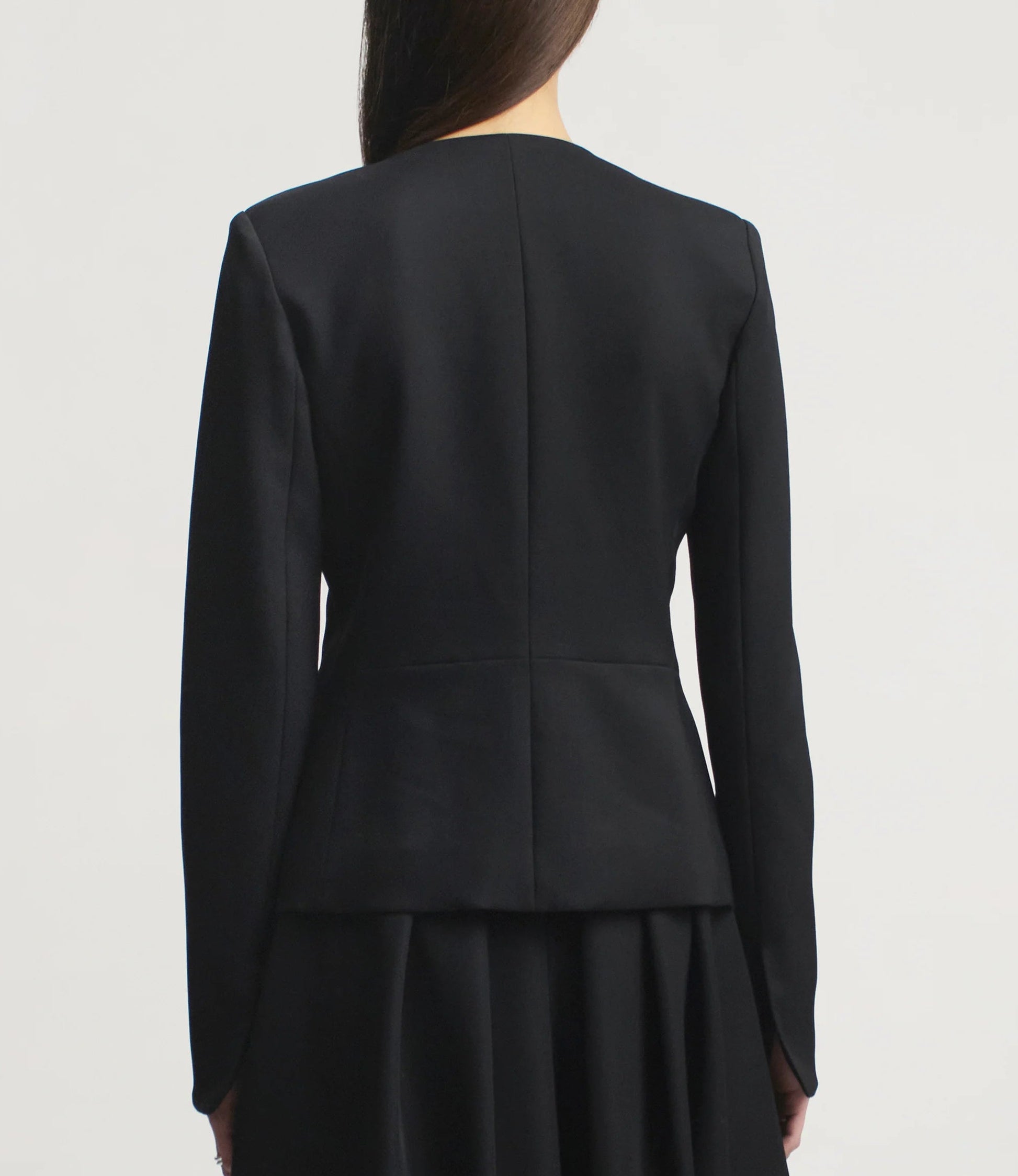 A woman with straight brown hair is shown from behind, wearing the Kallmeyer Tilda Jacket and skirt in classic black Grampians Twill against a plain light background.