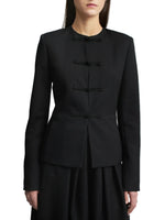 A woman stands against a plain white background, wearing the Kallmeyer Tilda Jacket—a black, collarless slim style featuring handmade frog closures—paired with a skirt. Her face is partially out of frame.