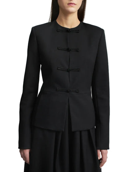 A woman stands against a plain white background, wearing the Kallmeyer Tilda Jacket—a black, collarless slim style featuring handmade frog closures—paired with a skirt. Her face is partially out of frame.