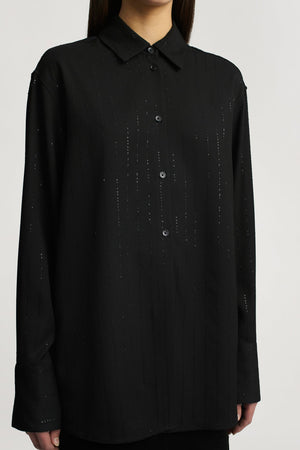 A person wearing the Kallmeyer Mollie Button Down, a relaxed fit black shirt with subtle vertical pinstripes, stands against a plain background.