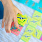A hand holds a Giant Shiny Dinosaur Dominos card by Eeboo, featuring colorful dinosaur art and holographic foil, over a surface scattered with more blue, yellow, and green dino dominos.