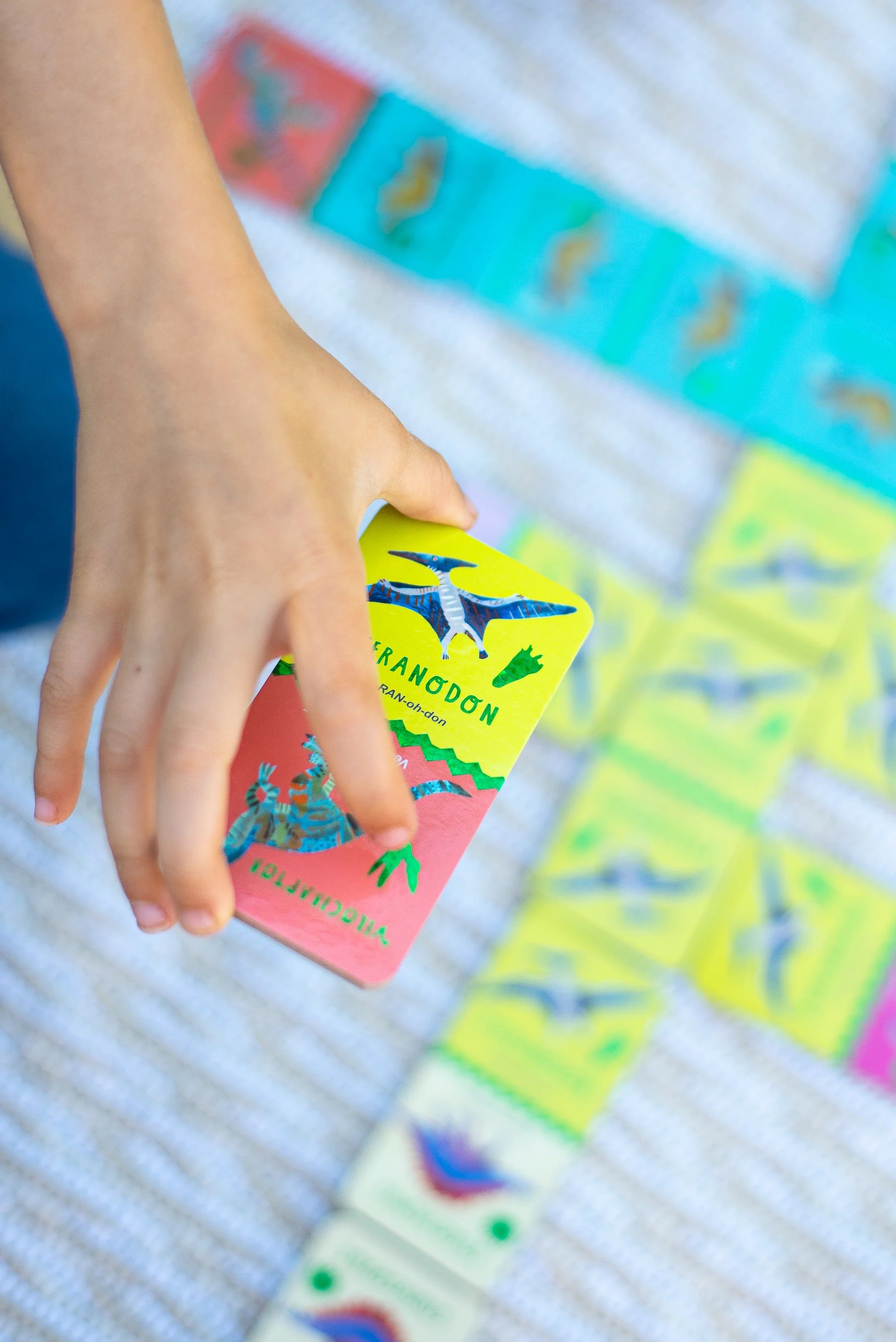 A hand holds a Giant Shiny Dinosaur Dominos card by Eeboo, featuring colorful dinosaur art and holographic foil, over a surface scattered with more blue, yellow, and green dino dominos.