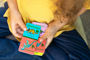 A child, wearing a yellow shirt and blue pants, plays with Eeboo’s Giant Shiny Dinosaur Dominos—colorful cards featuring illustrated dinosaurs and their names, all accented with shiny holographic foil for an extra-special learning experience.