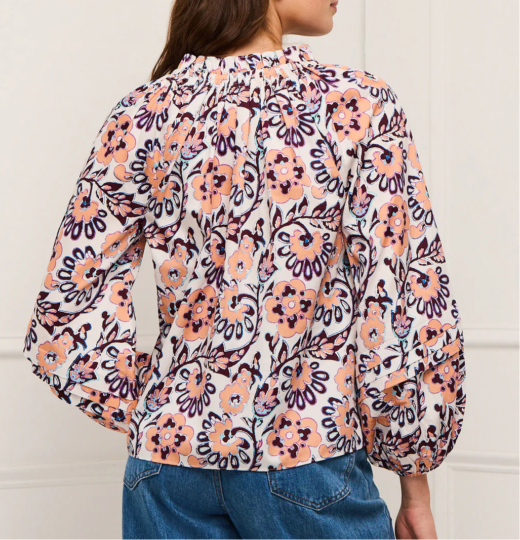 A woman is shown from the back wearing the Figue Donna Top, a long-sleeve blouse with a peach, purple, and white floral print and blouson sleeves, paired with blue jeans.