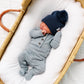 A newborn baby wearing the Lou Lou & Company Baby Boys’ and Boys’ Miller Plush Knit Top & Bottoms in grey, featuring built-in footies and a fold-over waistband crafted from silky-soft knit fabric, lies in a wicker bassinet lined with a white cushion.