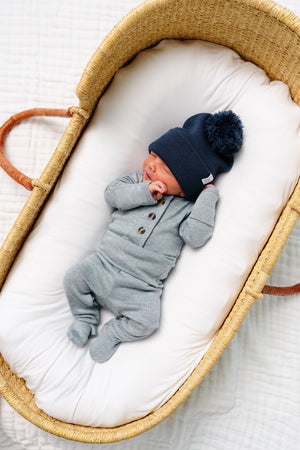 A newborn baby wearing the Lou Lou & Company Baby Boys’ and Boys’ Miller Plush Knit Top & Bottoms in grey, featuring built-in footies and a fold-over waistband crafted from silky-soft knit fabric, lies in a wicker bassinet lined with a white cushion.