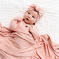 Nestled in the silky-soft fabric of the Lou Lou & Company Baby Girls’ and Girls’ Piper Plush Knit Top & Bottoms, a baby wrapped in cozy pink knitwear with a matching bow headband lies on a white quilted surface, looking up contentedly.