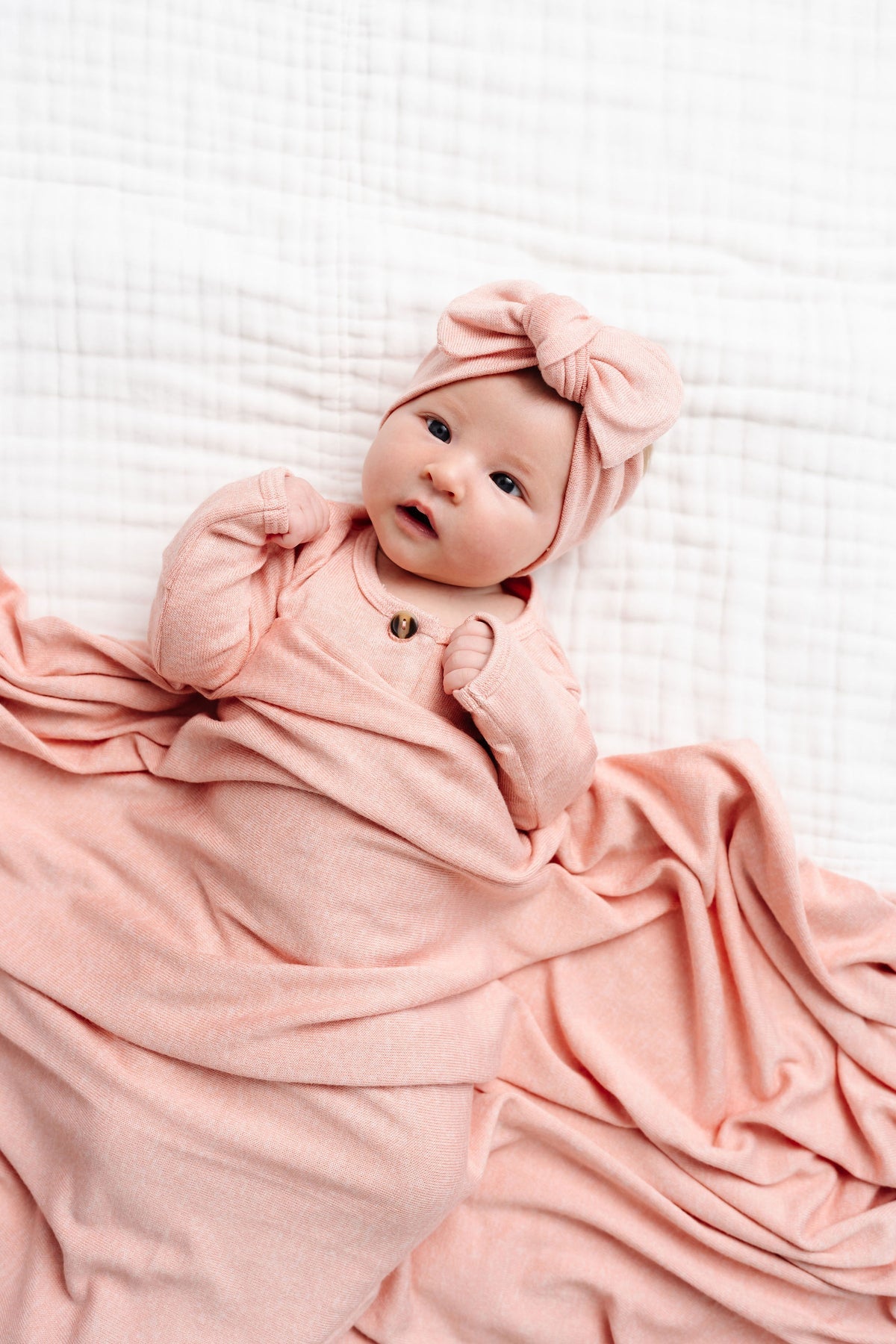 Nestled in the silky-soft fabric of the Lou Lou & Company Baby Girls’ and Girls’ Piper Plush Knit Top & Bottoms, a baby wrapped in cozy pink knitwear with a matching bow headband lies on a white quilted surface, looking up contentedly.