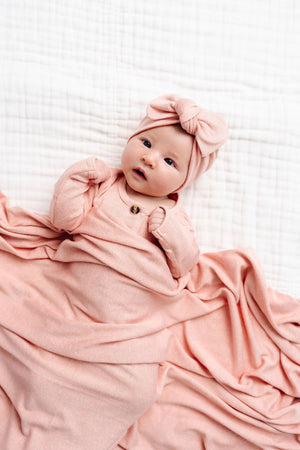 Nestled in the silky-soft fabric of the Lou Lou & Company Baby Girls’ and Girls’ Piper Plush Knit Top & Bottoms, a baby wrapped in cozy pink knitwear with a matching bow headband lies on a white quilted surface, looking up contentedly.