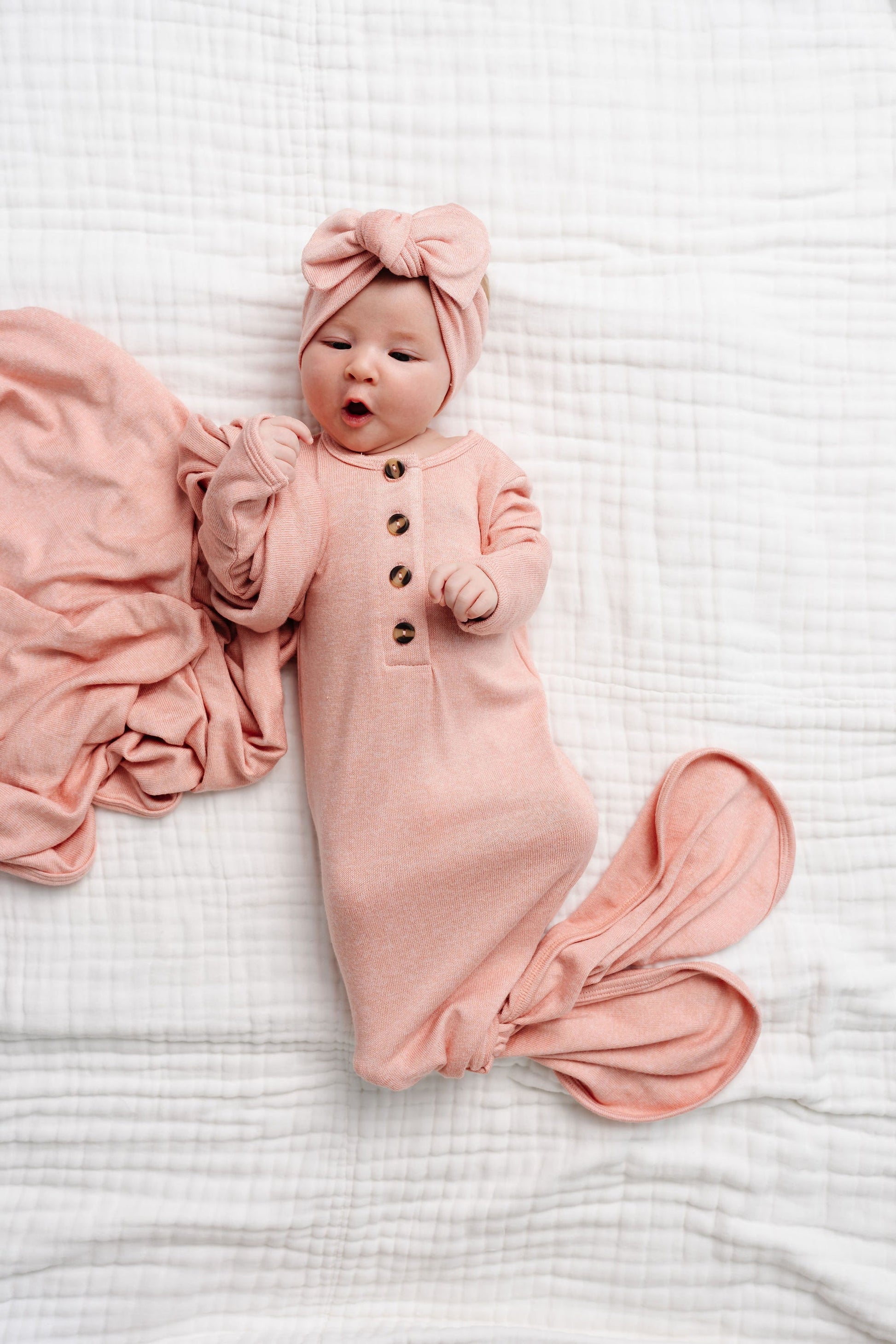 Lou Lou & Company’s Baby Girls’ Piper Plush Knit Knotted Gown features a soft pink design with a matching headband, shown here on a white quilted blanket with a pink blanket nearby—perfect for hassle-free diaper changes.