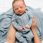 A newborn baby sleeps peacefully in an adult’s arms, dressed in a cozy Lou Lou & Company Baby Boys’ Miller Plush Knit Zipper Footie in light blue, with a silky-soft blue blanket underneath.