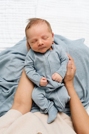 A newborn baby sleeps peacefully in an adult’s arms, dressed in a cozy Lou Lou & Company Baby Boys’ Miller Plush Knit Zipper Footie in light blue, with a silky-soft blue blanket underneath.