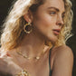 A woman with curly blonde hair wears elegant statement jewelry—a gold floral necklace, earrings, and the Anabel Aram Wildflowers Gold Wide Cuff—resting her hand on her shoulder against a natural outdoor background.