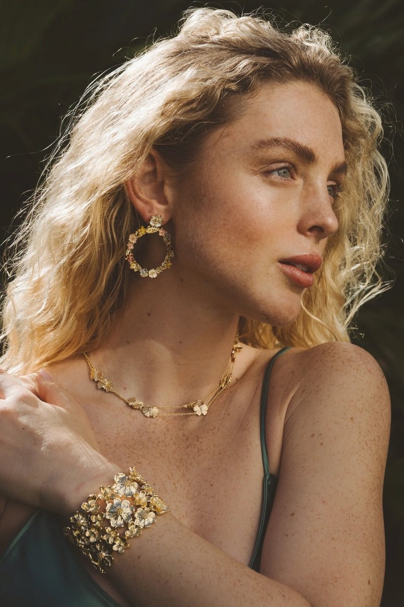 A woman with curly blonde hair wears elegant statement jewelry—a gold floral necklace, earrings, and the Anabel Aram Wildflowers Gold Wide Cuff—resting her hand on her shoulder against a natural outdoor background.