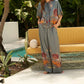 Wearing Carolina K Palazzo Pants, a person in a gray outfit with colorful patterns stands by a mustard-yellow couch near a pool, surrounded by lush green plants.