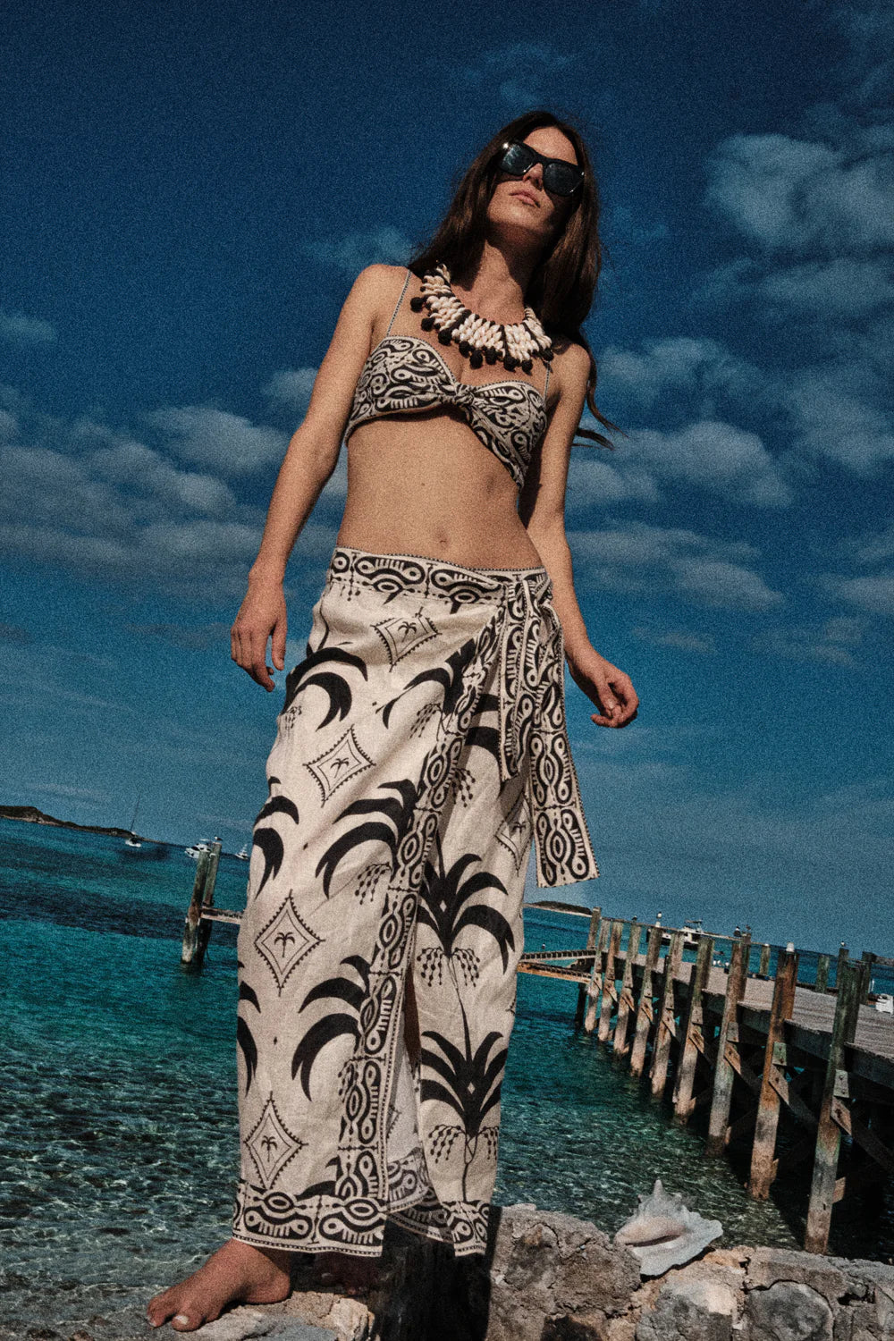 A woman stands barefoot on rocks by the ocean in the Johanna Ortiz Significado Poderoso Ankle Skirt, with a dock and clear blue water behind her under a partly cloudy sky.