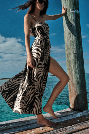 Woman in Johanna Ortiz’s Palms Nuevo Destino Midi Skirt stands barefoot on a dock by turquoise water, sunglasses on and wind-blown hair—effortless resort style in this chic zebra-print piece.