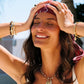 A person wears an 18K gold plated Milos Stone Fish Charm necklace featuring a small shell and a cylindrical stone charm, complemented by layered bracelets on their wrists.