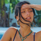 A woman wears a layered turquoise beaded Milos Stone Fish Charm necklace by Anabel Aram, featuring an 18K gold-plated fish with a dangling green stone pendant.