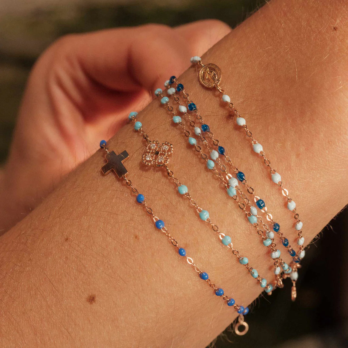 A wrist featuring the timeless design of a Gigi Clozeau Madone Charm Bracelet 6.7" adorned with elegant resin pearls, small decorative pendants, and beads in various blue and gold shades.