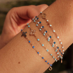 A wrist adorned with four delicate bracelets in blue and gold shades, featuring cross, flower, and circular charms. The Gigi Clozeau Madone Charm Bracelet 6.7" adds elegance with its 18-carat gold accents and refined resin pearls.