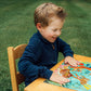 A child with light brown hair enthusiastically assembles the Eeboo Deep Sea Treasure 20 Piece Puzzle at a wooden table outdoors on a grassy lawn.