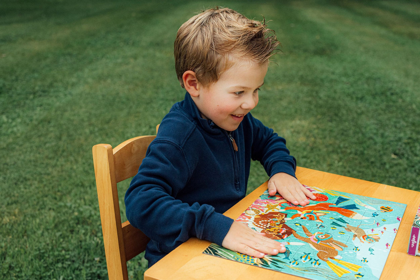 A boy in a blue sweater sits at a wooden table outdoors, engrossed in the eeboo Deep Sea Treasure 20 Piece Puzzle by Eeboo, featuring jumbo-sized pieces.